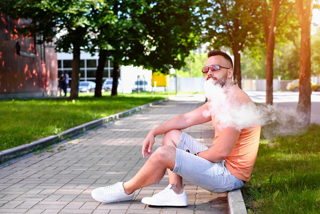 Man sitting down by a path blowing out a cloud of vapour.