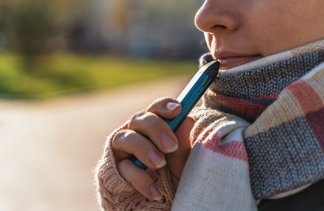 Woman wearing a warm scarf vaping a pod device suitable for using with nic salts.