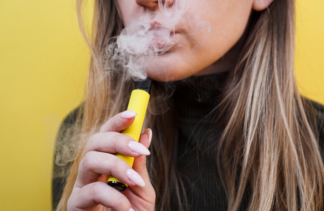 A woman holds a yellow disposable vape device while exhaling vapour.